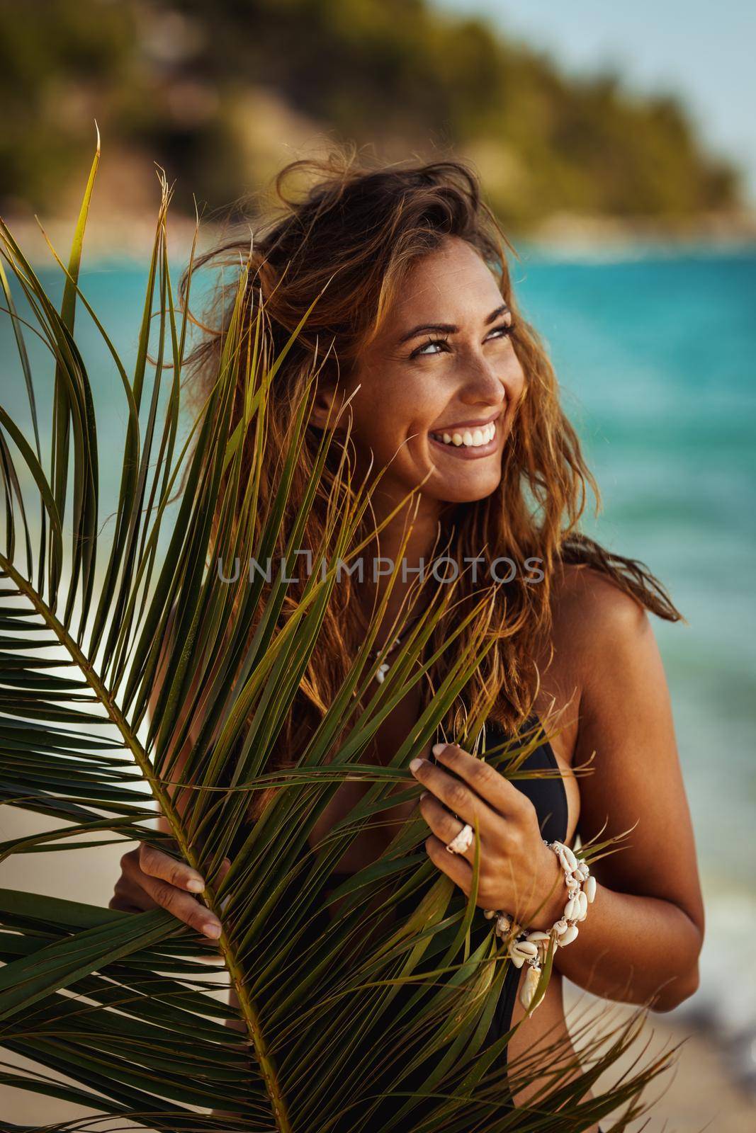 Portrait of an attractive young woman is posing and enjoying covered with palm tree leaf at the tropical beach.