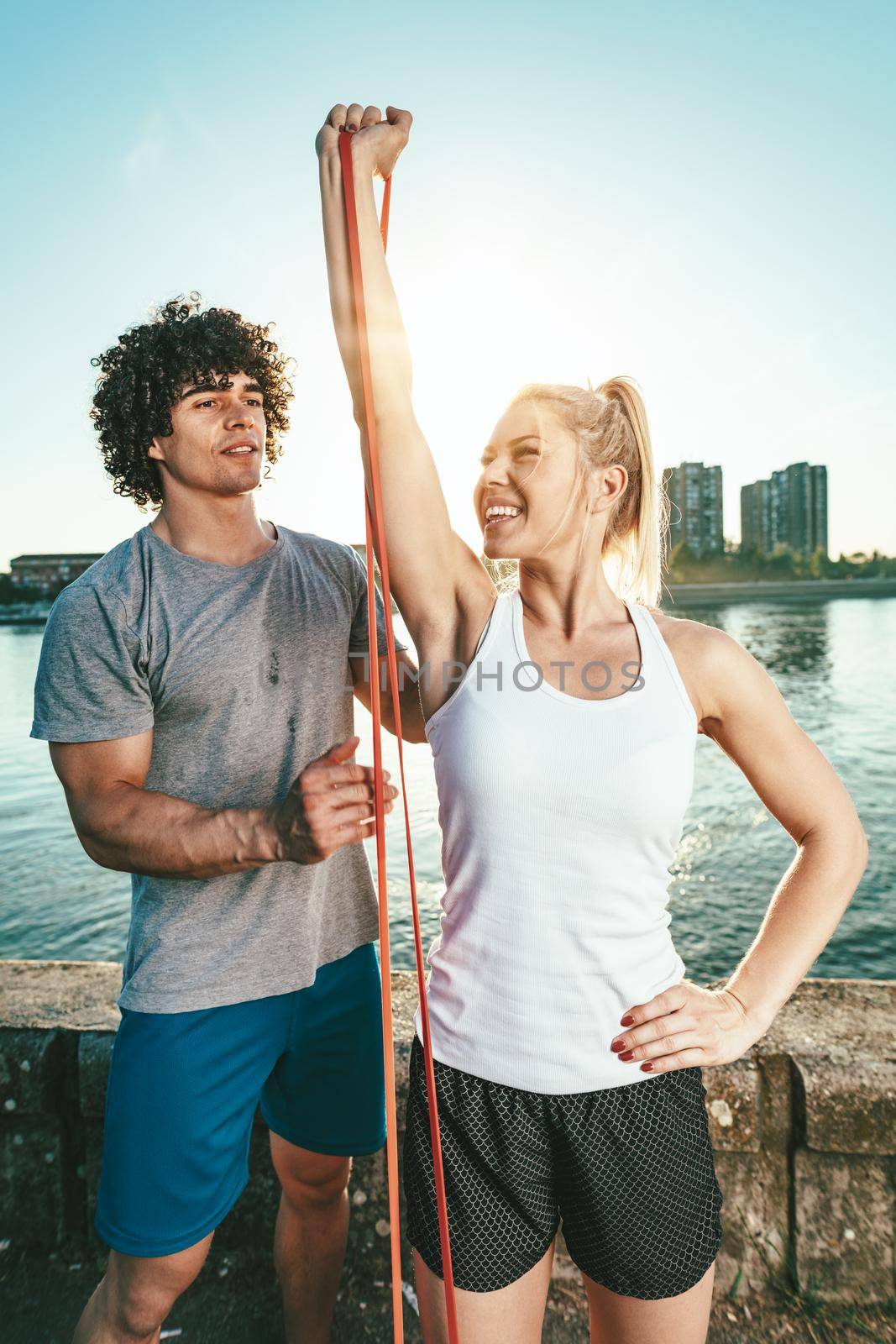 Young fitness couple is doing workout with rubber band by the river in a sunset. The woman is stretching arms and the man supports her.