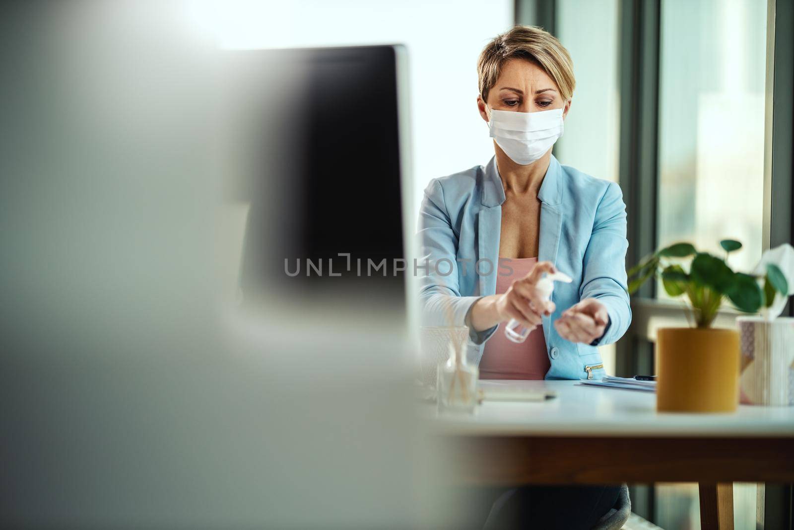 Business woman in a medical protective mask uses antibacterial antiseptic gel for hands disinfection and works from home at the computer during self-isolation and quarantine to avoid infection during flu virus outbreak and coronavirus epidemic.