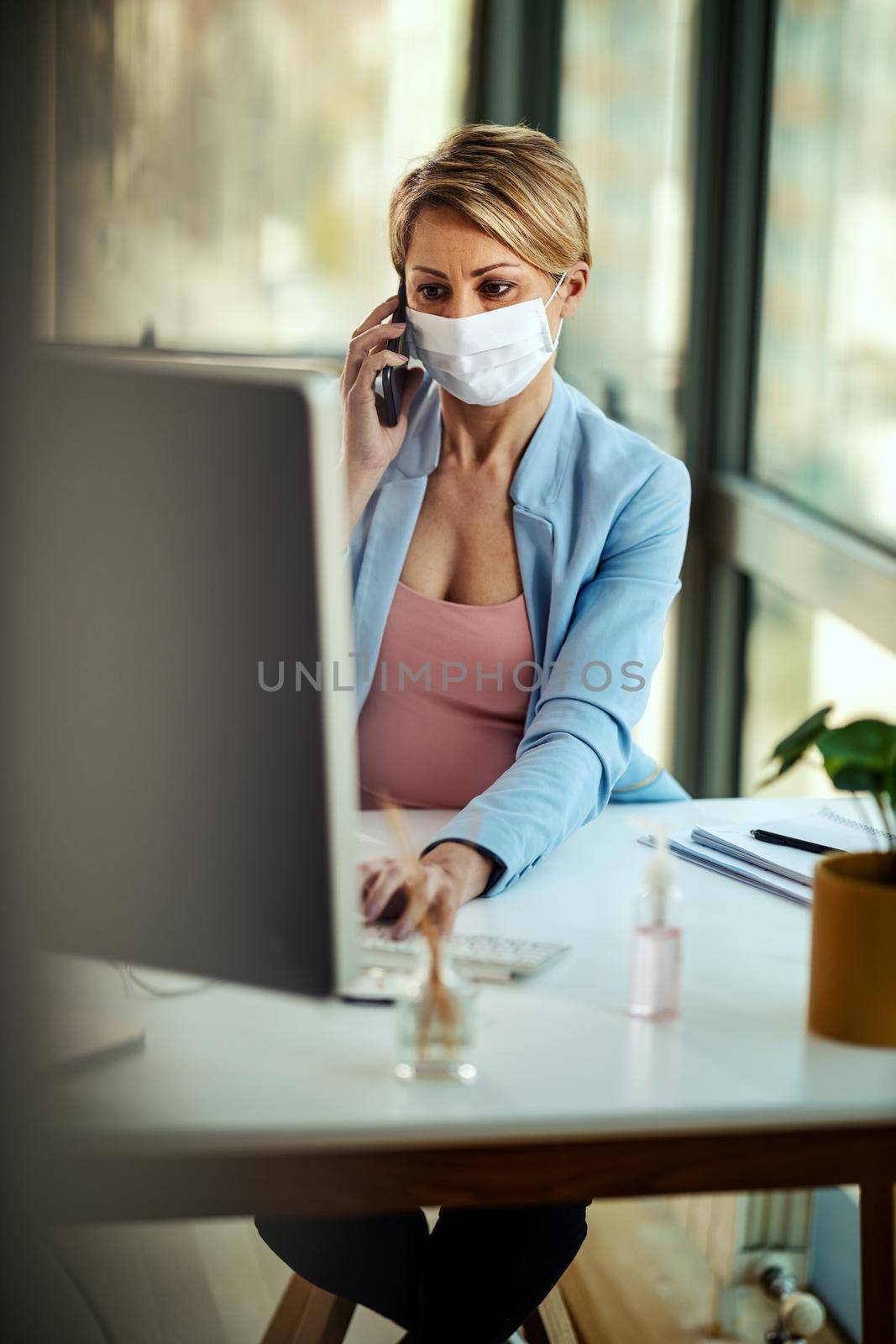 Business woman in a medical protective mask works at the computer and talking on a smartphone during self-isolation and quarantine to avoid infection during flu virus outbreak and coronavirus epidemic.