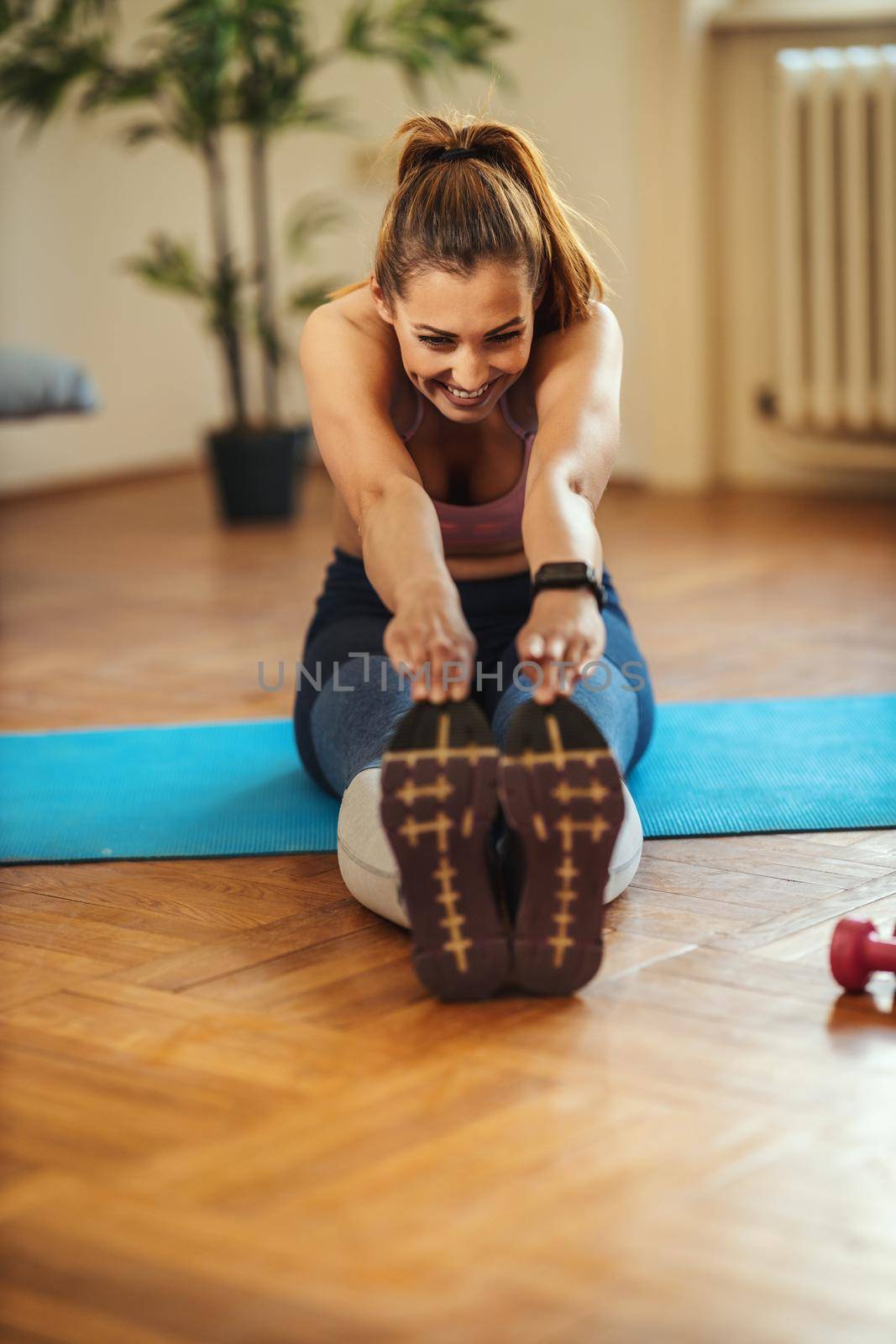 Young smiling woman is doing stretching exercises in the living room on floor mat at home in morning sunshine.