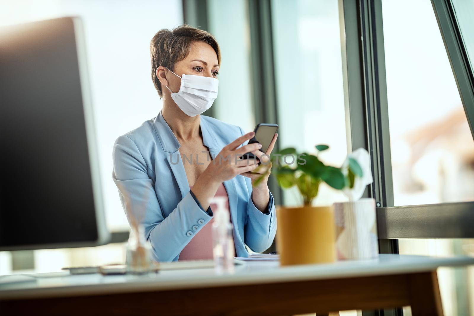 Business woman in a medical protective mask works from home at the computer, using smartphone, during self-isolation and quarantine to avoid infection during flu virus outbreak and coronavirus epidemic.