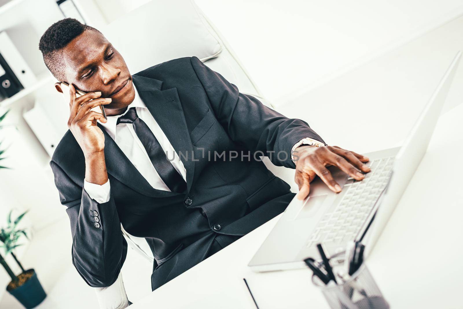 Pensive African businessman talking on smartphone and working on laptop in modern office.