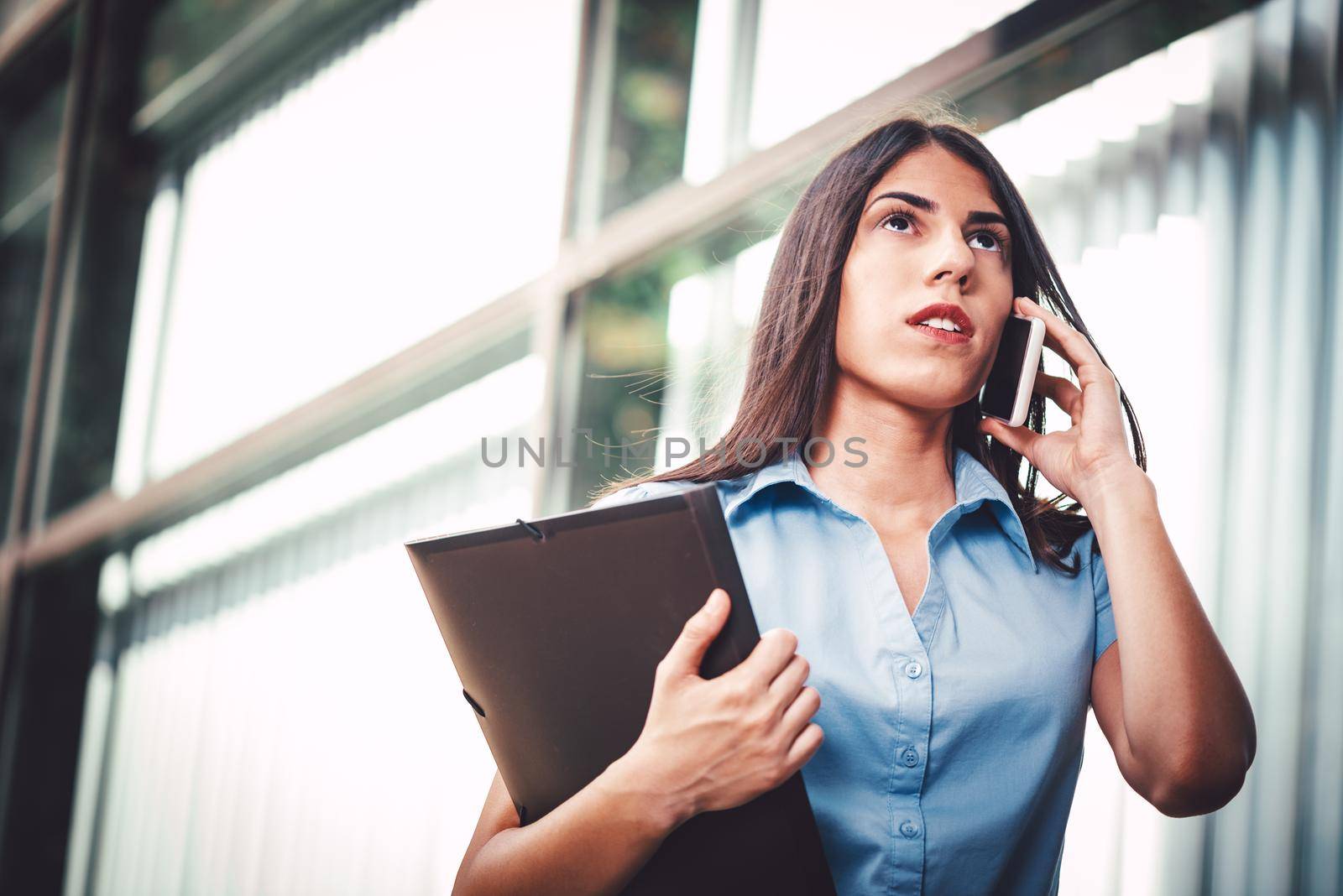 Emotional portrait of a pensive and beautiful young businesswoman talking on a smartphone and carries a file document on the background of a business center.