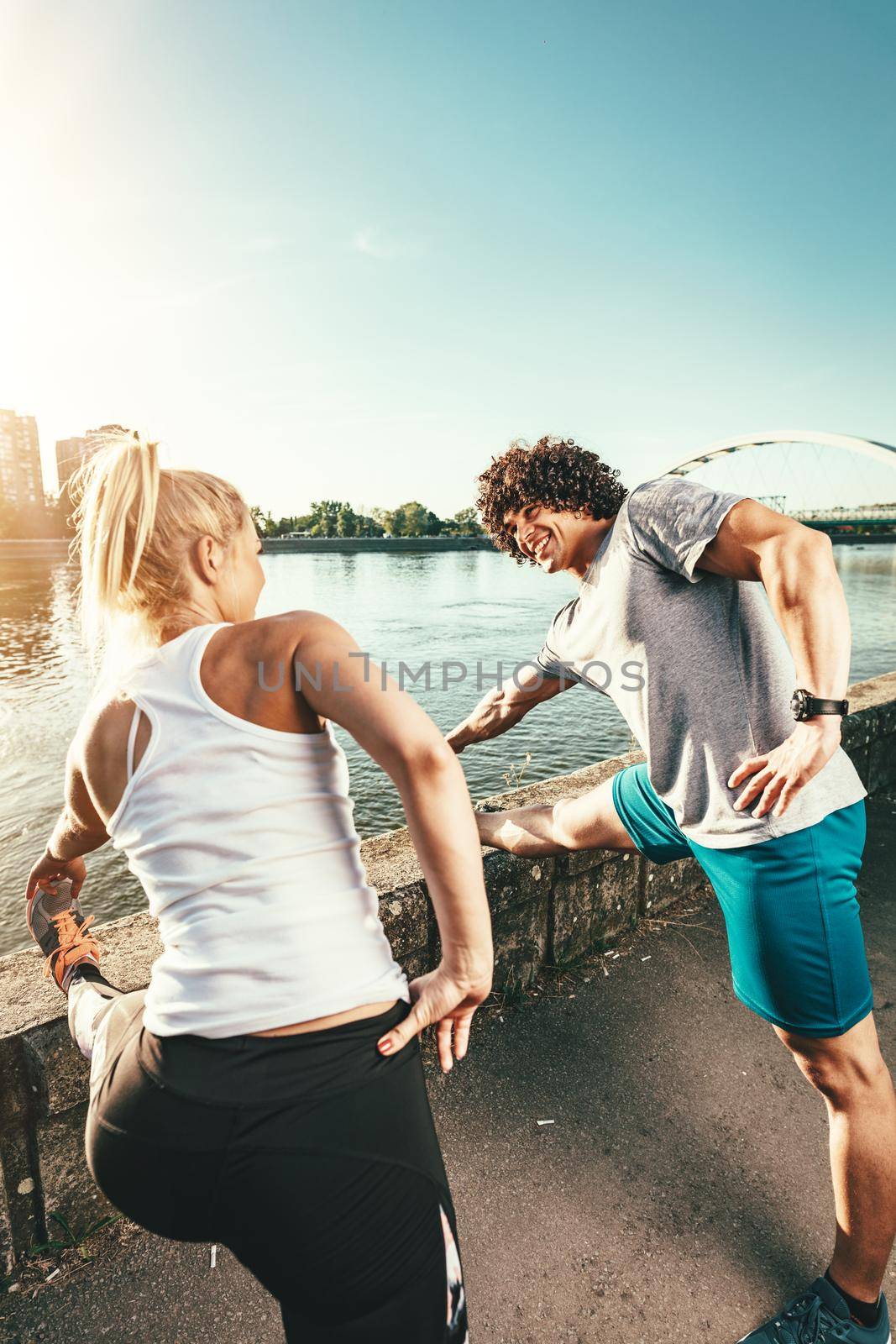 Young happy couple is training outdoors by the river, stretching legs by the wall at sunset.