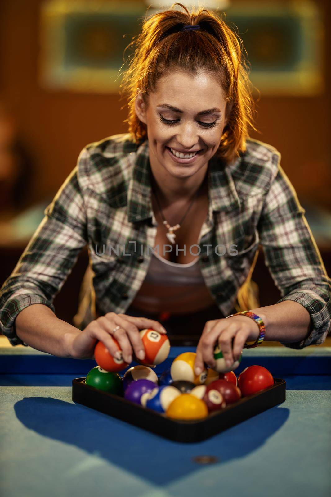 A young smiling woman places billiard balls on a pool table to play.