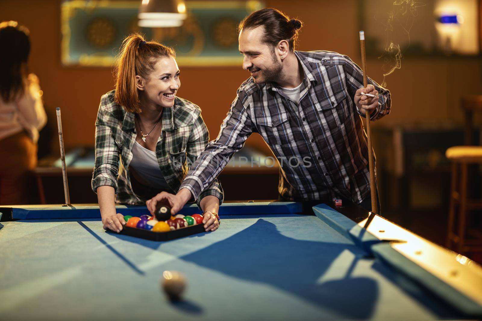Young smiling cheerful couple is playing billiards in bar after work. They are having fun and involved in recreational activity.