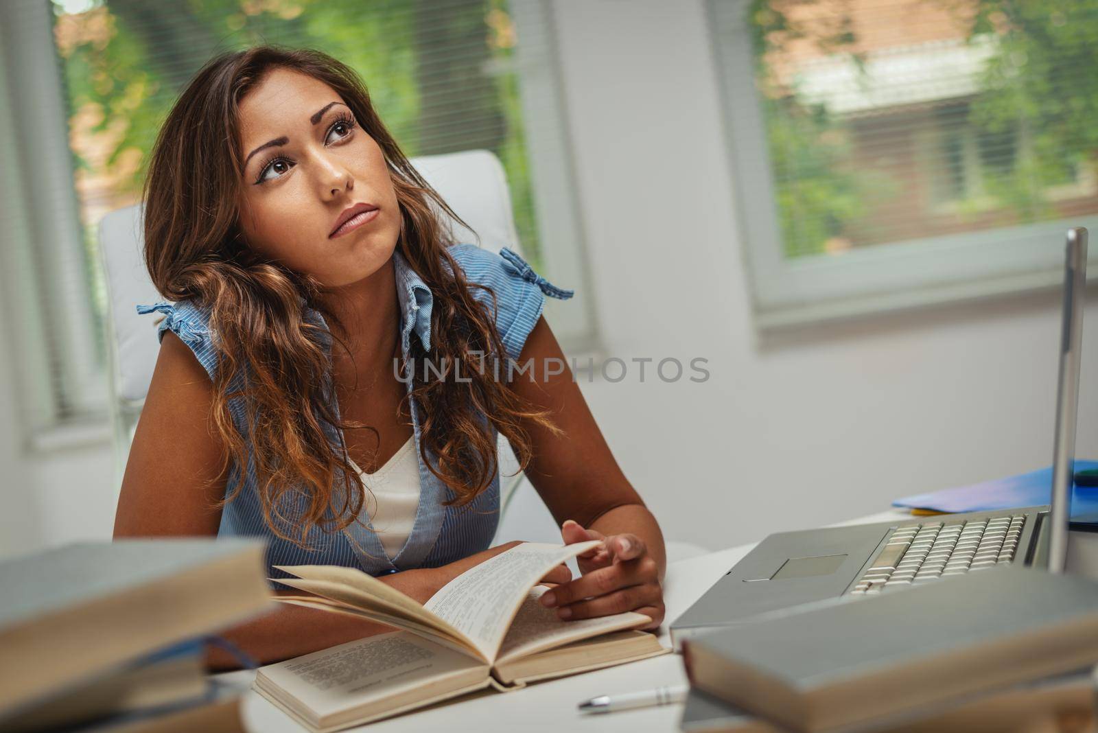 Beautiful pensive young female student  with book is preparing exam and learning lessons in school library.