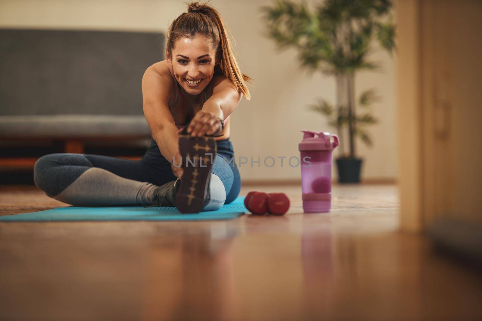 Young smiling woman is doing stretching exercises in the living room on floor mat at home in morning sunshine.