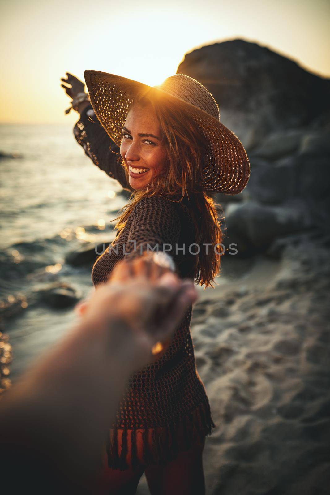 A beautiful young woman is having fun and relaxing on the beach at the sunset. She extends a hand to someone whose hand is seen.