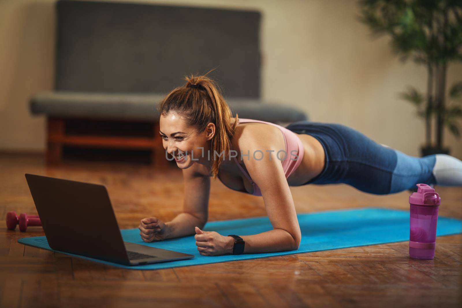 Young smiling woman is doing plank exercises in the living room on floor mat at home, looking at the laptop, in morning sunshine.