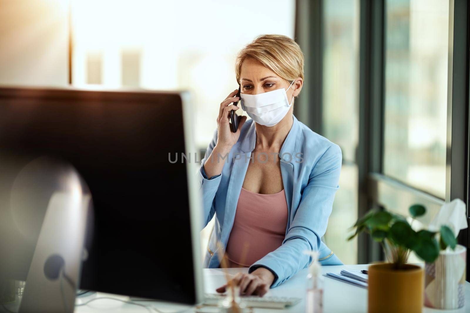 Business woman in a medical protective mask works at the computer and talking on a smartphone during self-isolation and quarantine to avoid infection during flu virus outbreak and coronavirus epidemic.