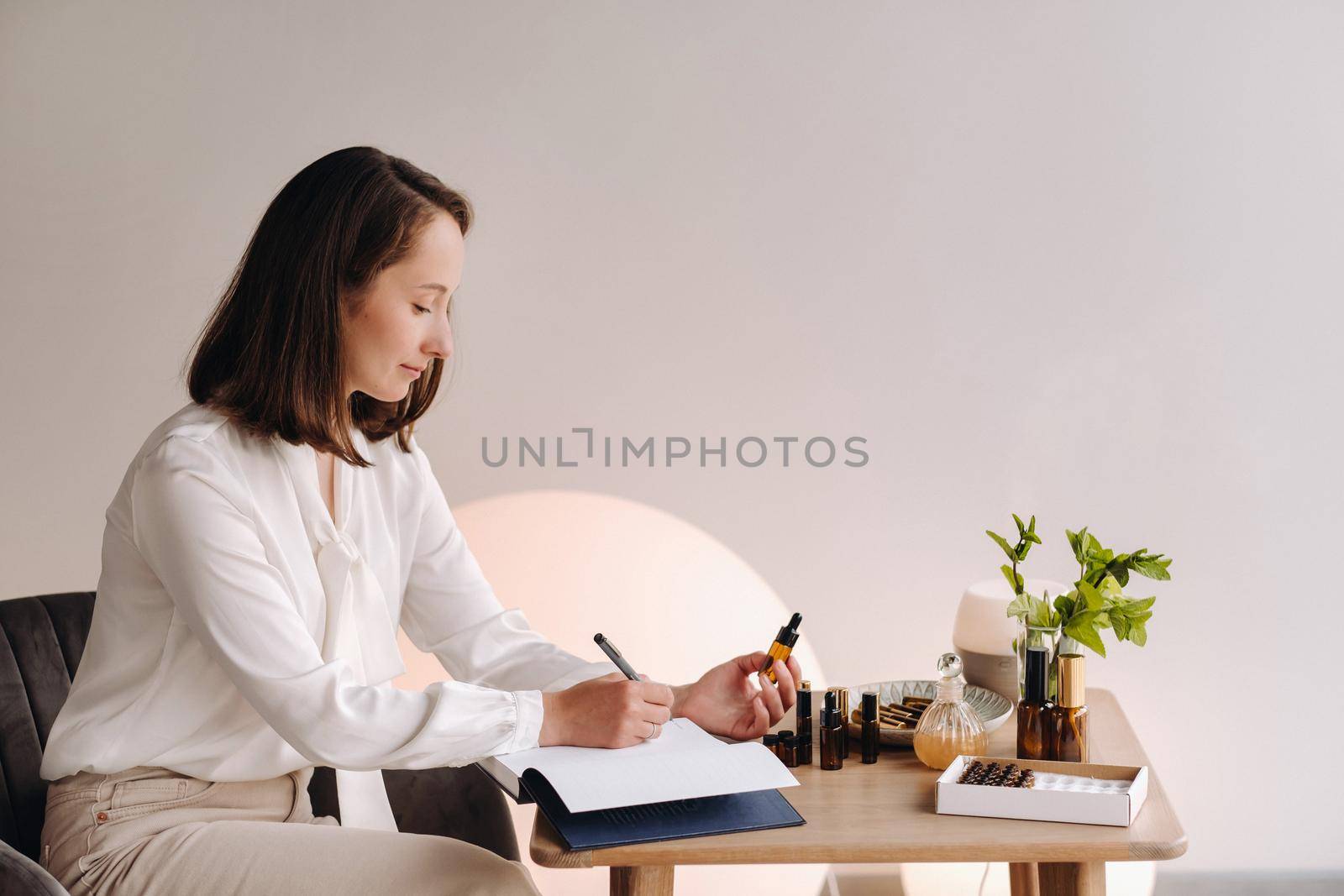 The aromatherapist girl is sitting in her office and holding a bottle of aromatic oil in her hands and writing something down. there are essential oils on the table by Lobachad