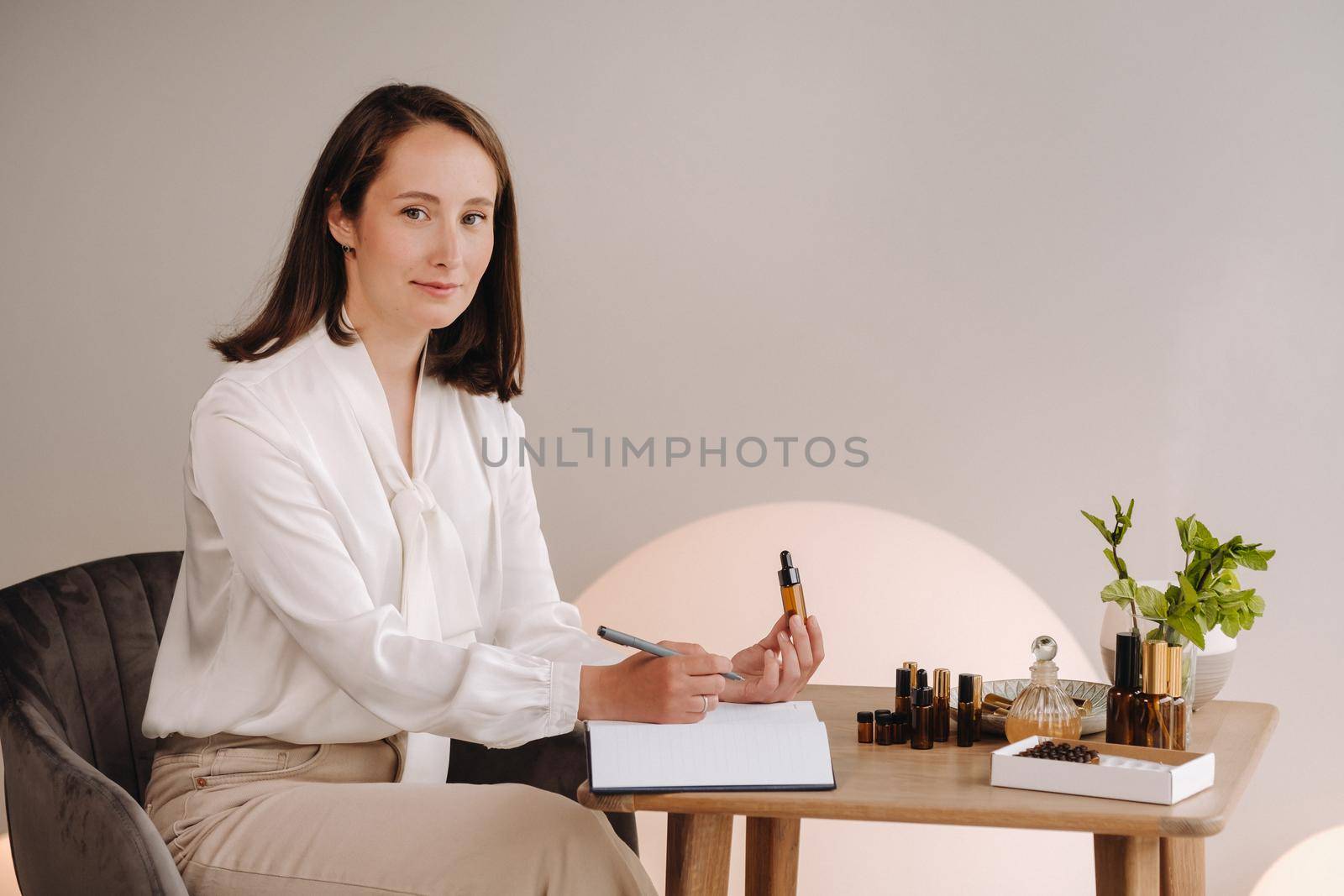 The aromatherapist girl is sitting in her office and holding a bottle of aromatic oil in her hands and writing something down. there are essential oils on the table.