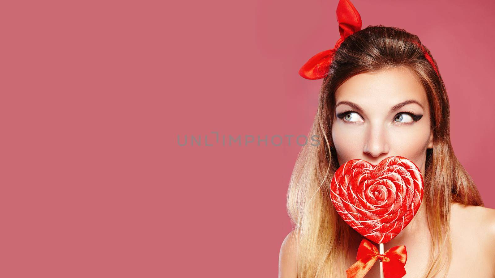 Sexy American Beauty. Pinup Girl with Red Bow Accessories in Blond Hair Staring to Copy Space Side. Playful Happy Woman with Sweet Candy Lollipop in Heart Symbol Shape. Beautiful Valentines Day Style