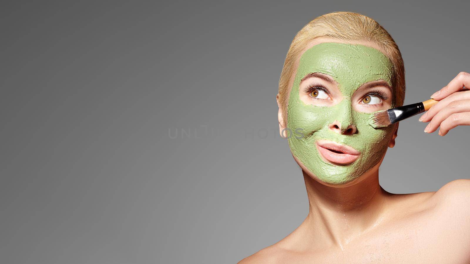 Beautiful Woman Applying Green Facial Mask. Beauty Treatments. Spa Girl Apply Clay Facial mask on grey background by MarinaFrost