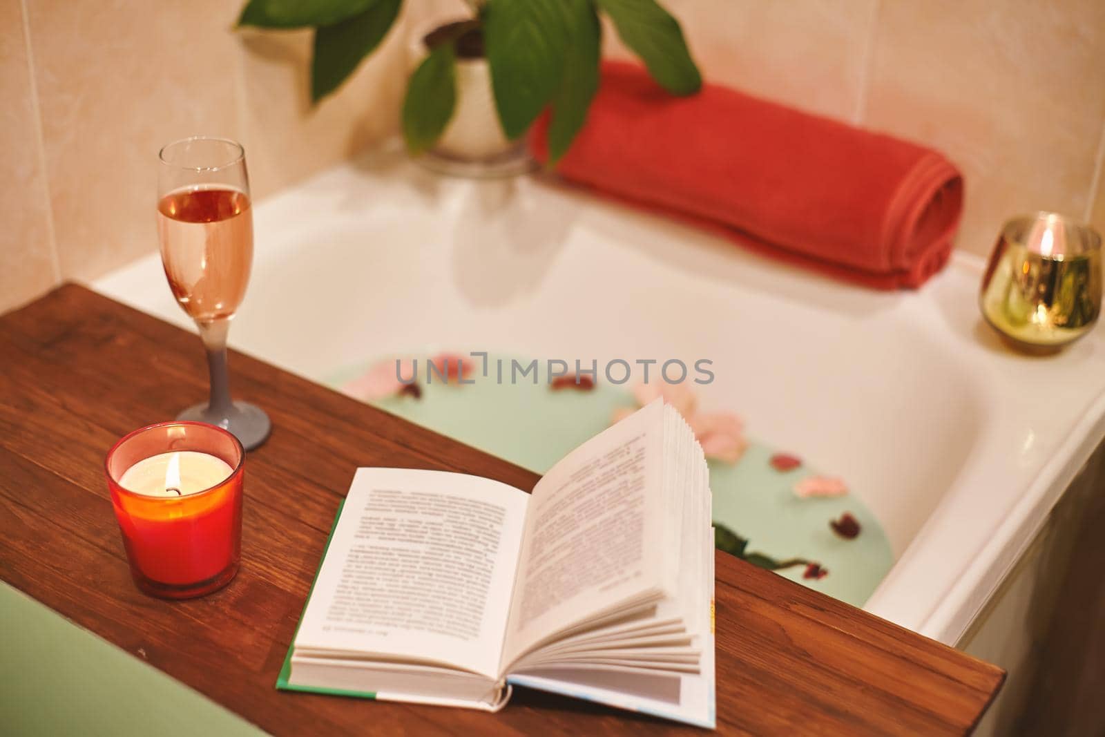 Bath with flower petals. Book, candles and glass of wine on a wood tray. Organic Spa Relaxation in comfort cozy bathroom by MarinaFrost