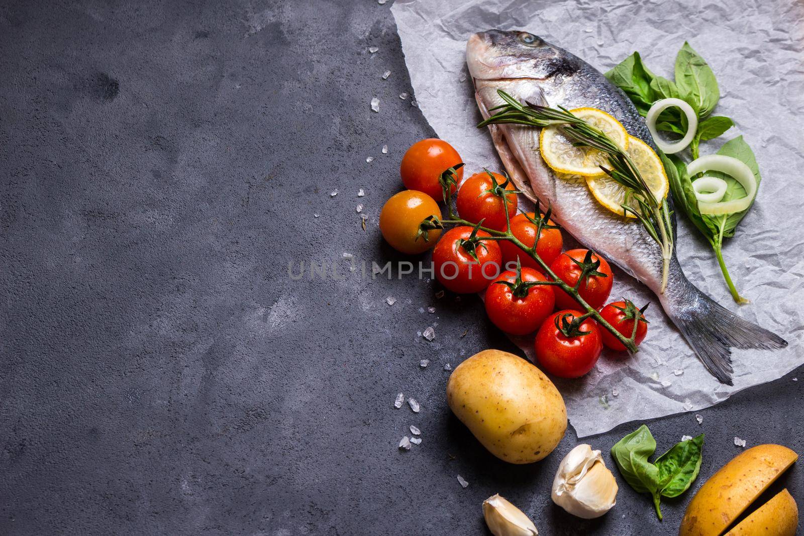 Raw fish with fresh ingredients ready to cook. Fish, lemon, herbs, potato, tomatoes. Ingredients for cooking on dark rustic background. Space for text. Diet and healthy food. Fish background. Top view