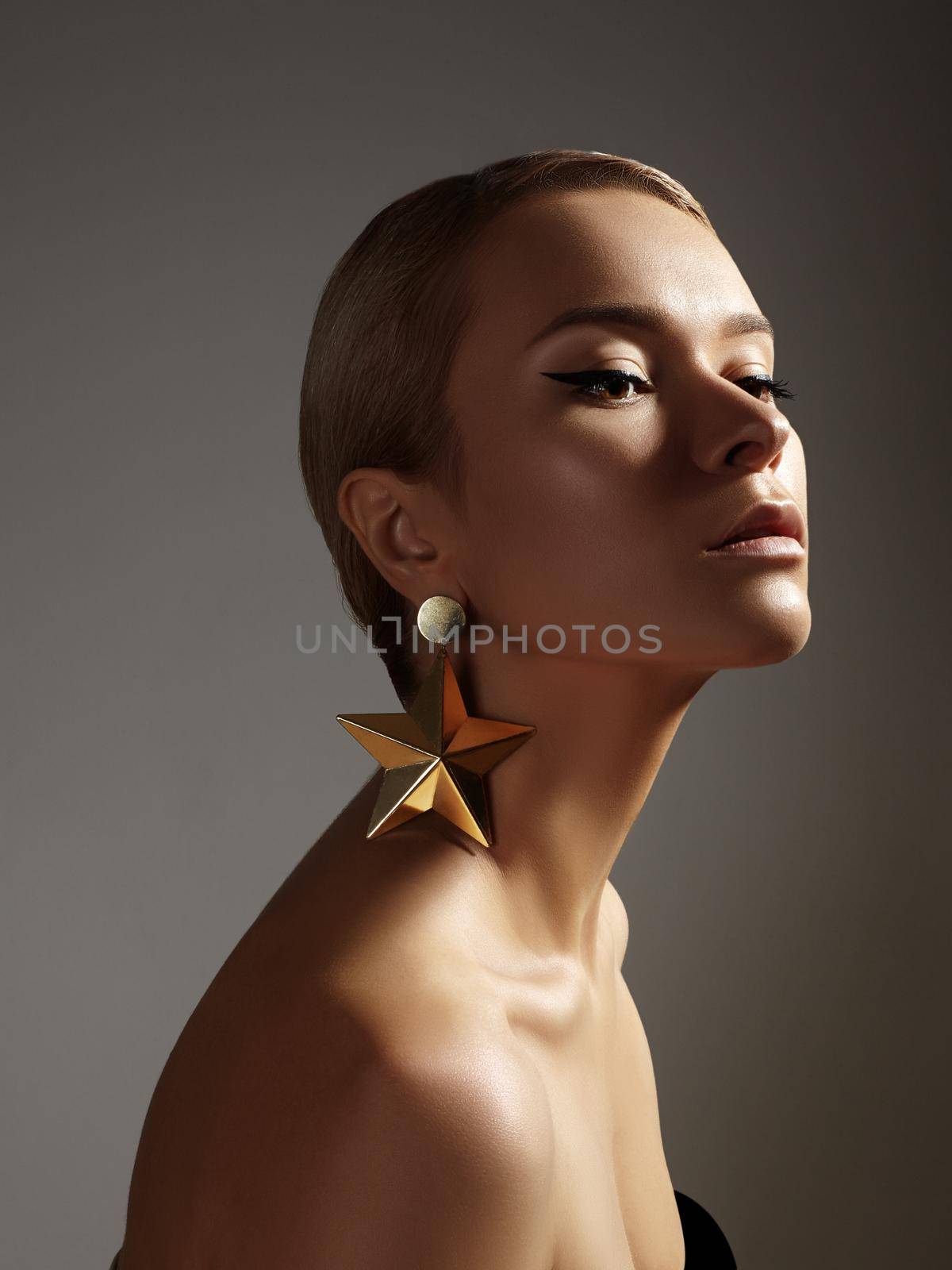 Beauty with Pure Skin on Grey Background with Luxury Accessories. Beautiful Woman Model in Femininity Pose with Fashion Celebratory Make-up, Sleek Hairstyle and Gold Earrings