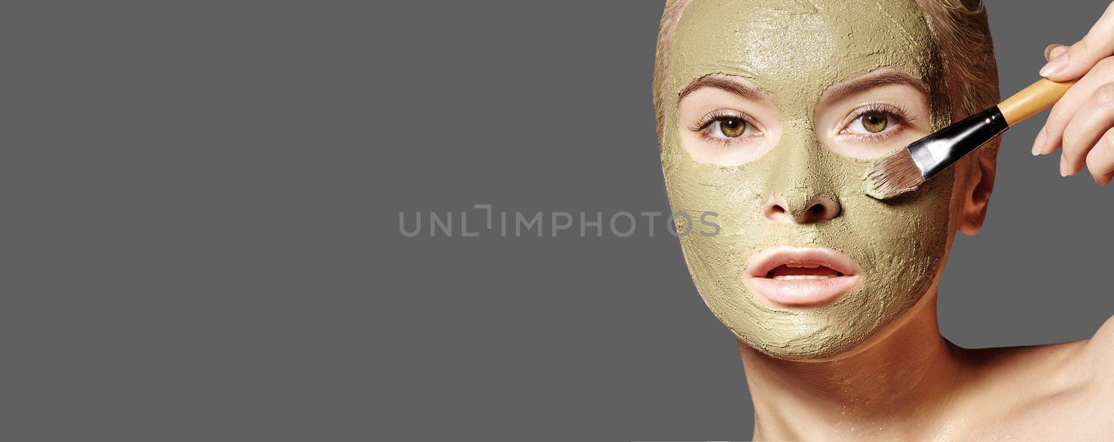 Beautiful Woman Applying Green Facial Mask. Beauty Treatments. Close-up Portrait of Spa Girl Apply Clay Facial mask on grey background.