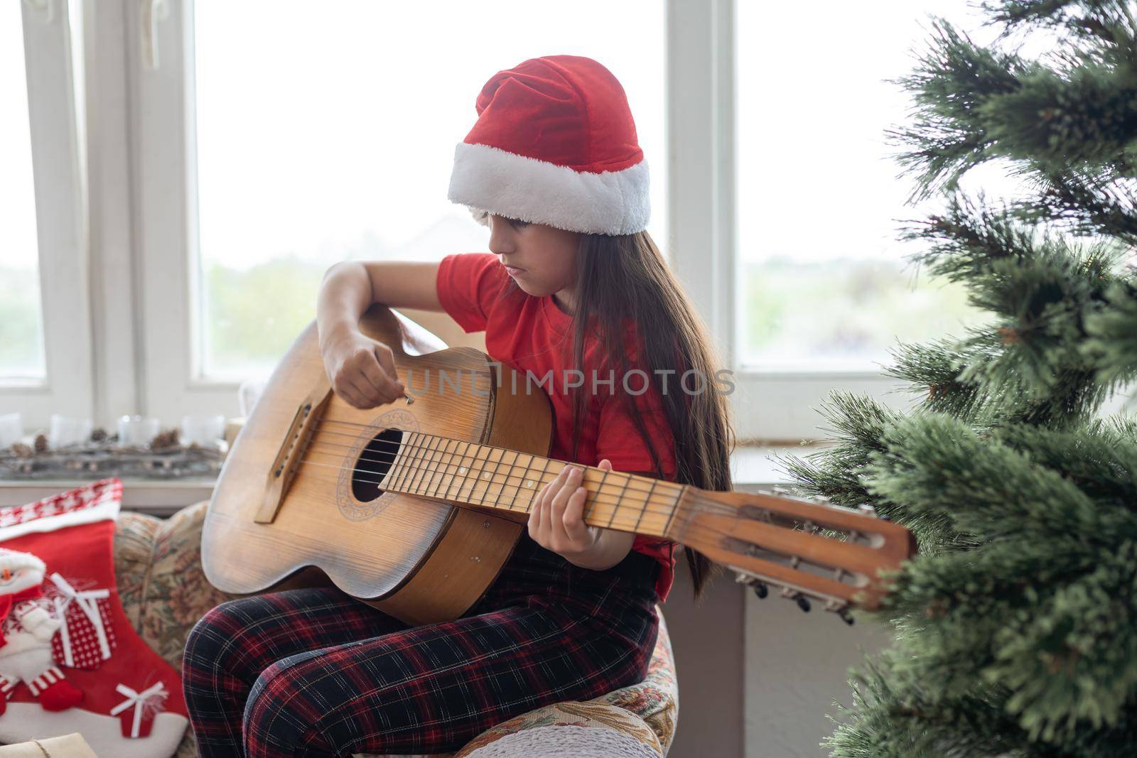Happy little girl in red hat playing the on the background of christmas tree. Talent kid having fun on winter holidays at cozy decorated home. by Andelov13