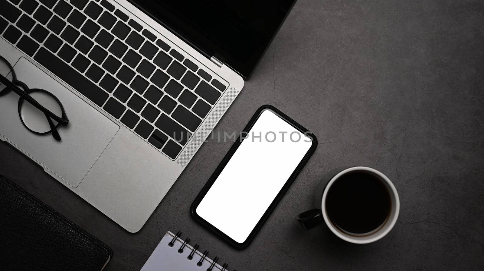 Mock up smart phone with blank screen, laptop computer, notebook and coffee cup on dark stone background. Flat lay.