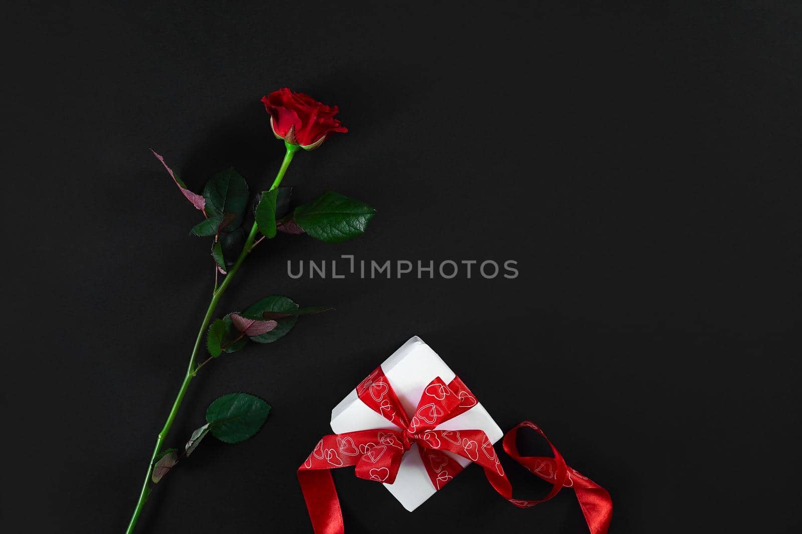 One dark red rose with red ribbon and gift box on black background. Romantic Valentines holidays concept. Valentine's day greeting card. Top view. Copy space. Still life. Flat lay