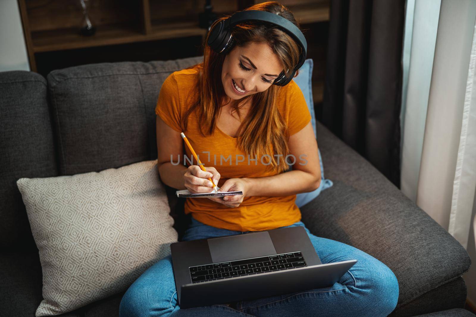 Attractive young woman sitting cross legged on the sofa and using her laptop and headphones to learning at home.