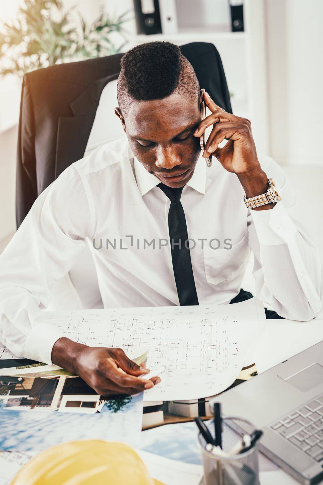 Pensive African architect talking on smartphone, analyzing blueprint and planning what to do next.