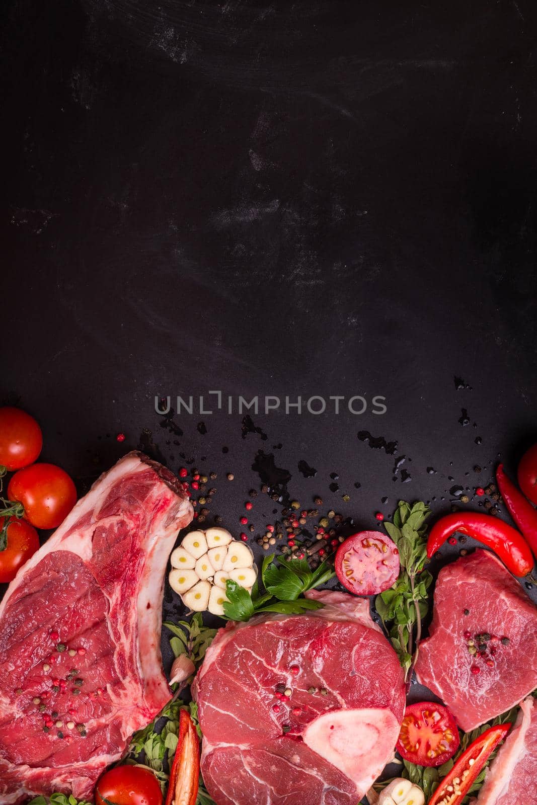 Raw juicy meat steaks ready for roasting on a black chalk board background. Rib eye steak on the bone, veal shank (ossobuco), fillet with cherry tomatoes, hot pepper and herbs. Space for text