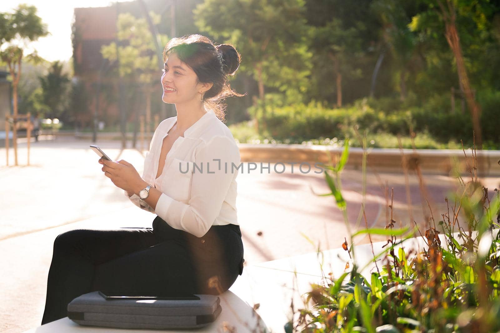smiling businesswoman checking her phone at sunrise sitting in a park in the financial district, concept of digital entrepreneur and urban lifestyle, copy space for text