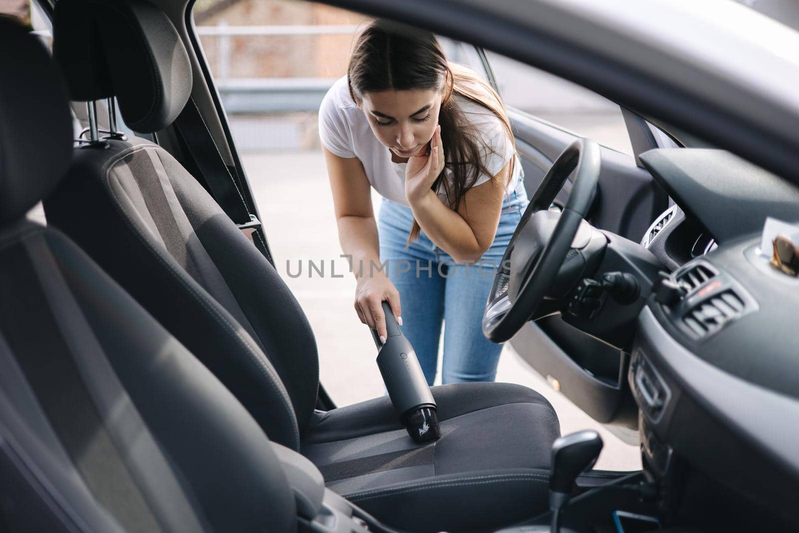 Attractive young woman using portable vacuum cleaner in her car. Woman vacuuming her car in the garage at home. Car interior cleaning.