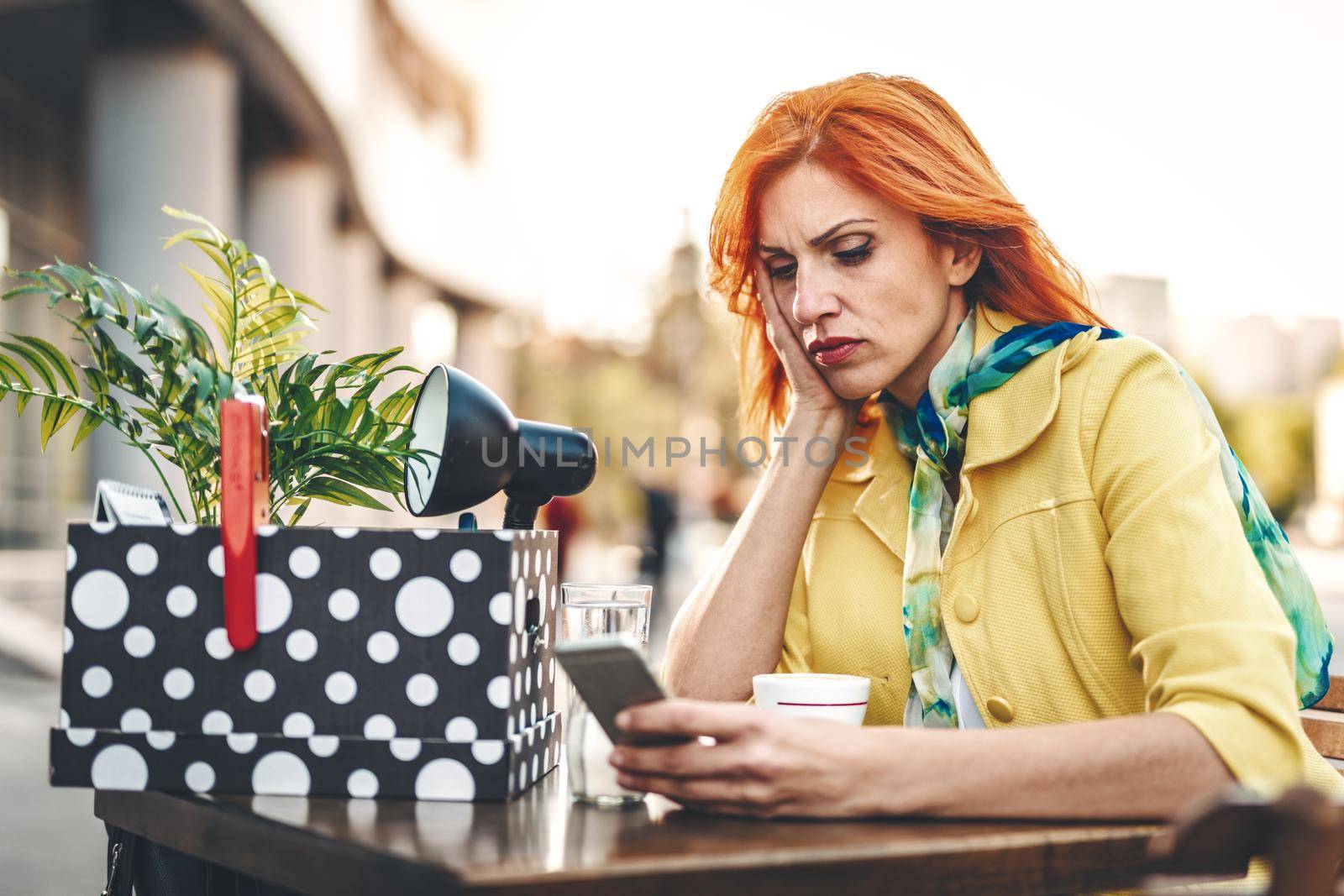 Serious pensive business woman is sitting in a street cafe with a box full of her personal belongings from the office, just after she got fired. 
