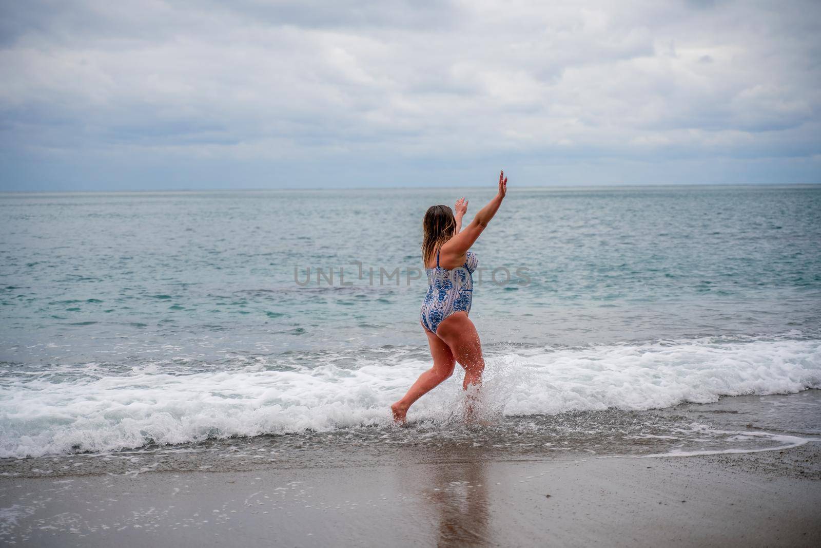 A plump woman in a bathing suit enters the water during the surf. Alone on the beach, Gray sky in the clouds, swimming in winter