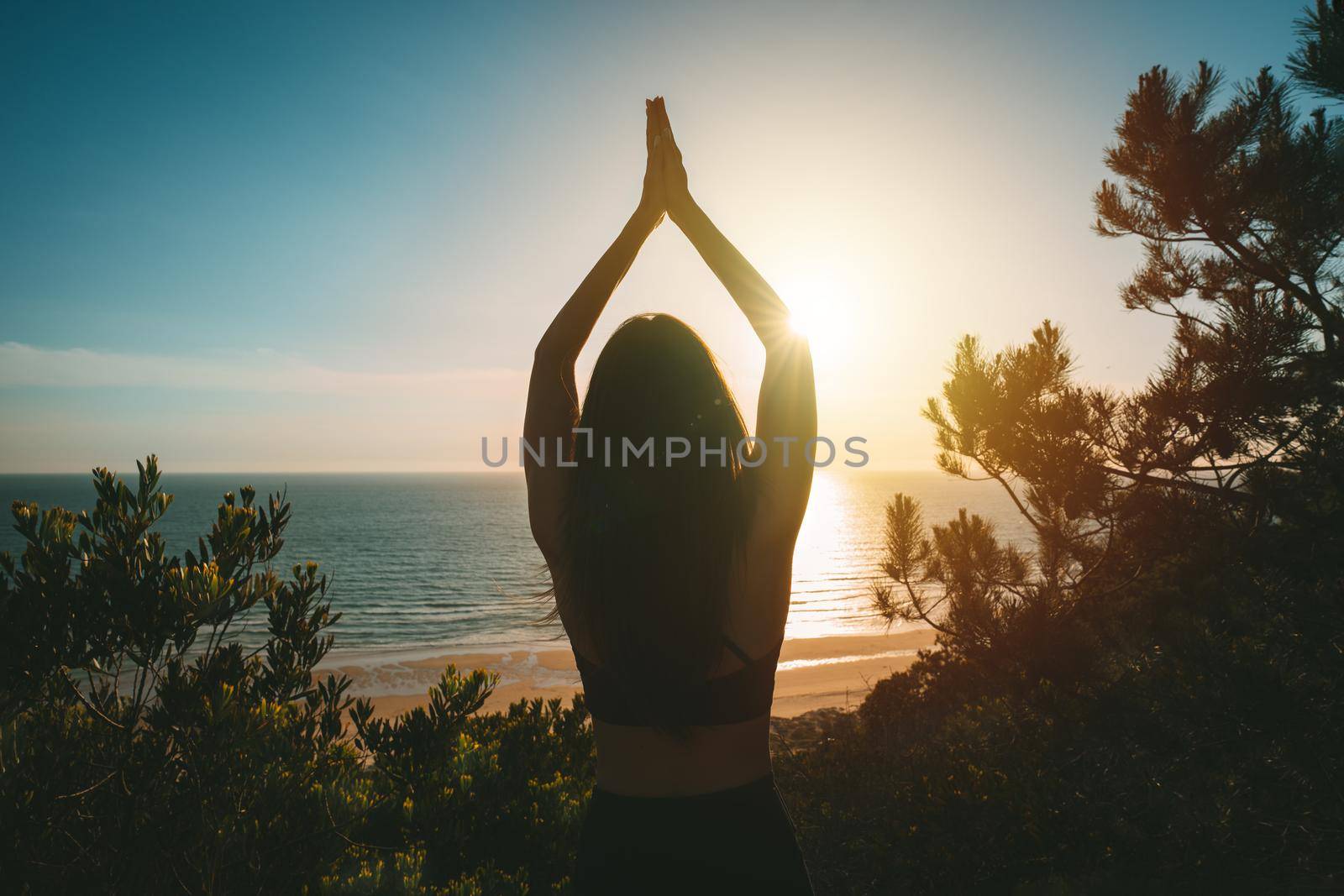 Silhouette of young healthy woman practicing yoga with ocean view at sunset. Rear view of woman standing with hands joined over head in tree pose. High quality photo