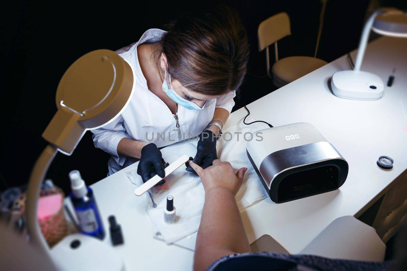 Cosmetologist in a protective mask, processing the nails on the client's hand by deandy