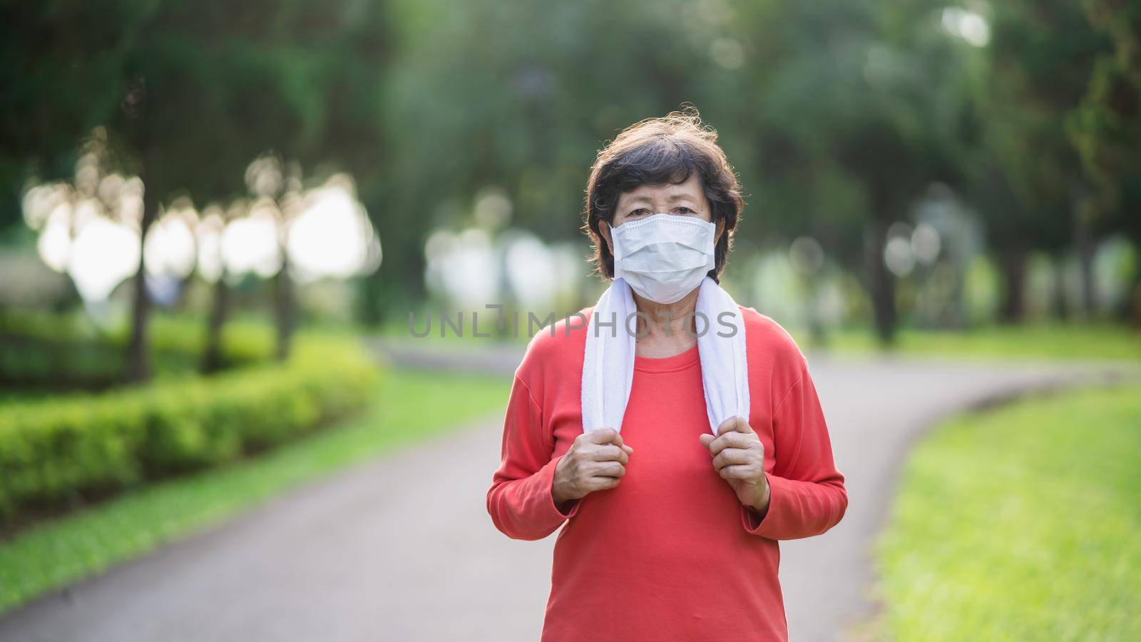 Athletic asian senior woman 60s wearing surgical mask and jogging. Beautiful senior asian woman running at the park on a sunny day.