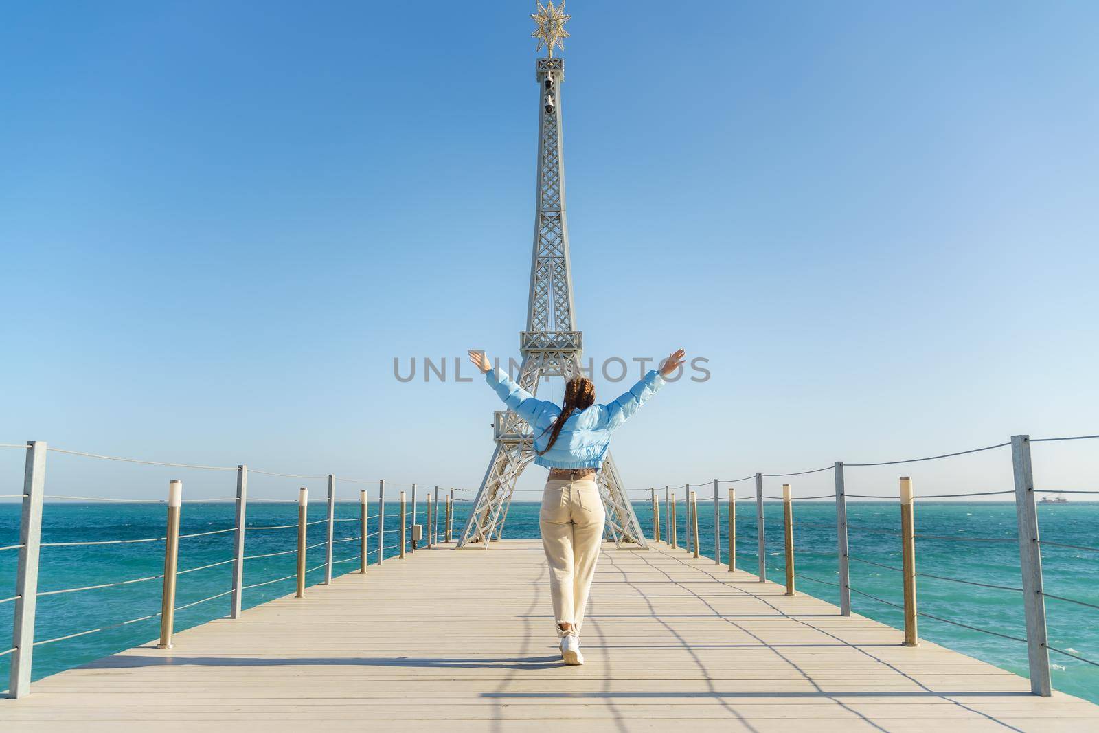 Large model of the Eiffel Tower on the beach. A woman walks along the pier towards the tower, wearing a blue jacket and white jeans. by Matiunina