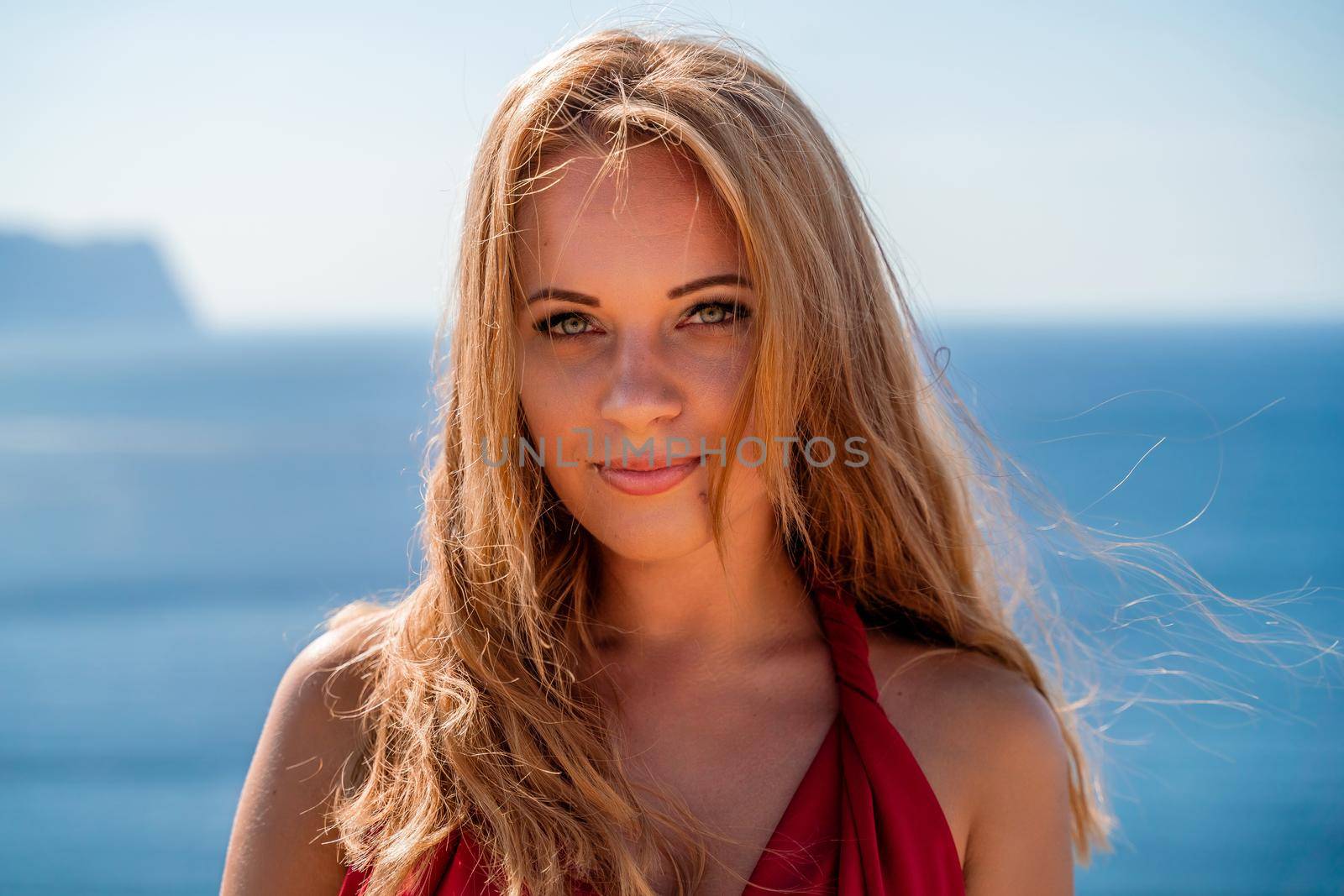 Smiling young woman in a red dress looks at the camera. A beautiful tanned girl enjoys her summer holidays at the sea. Portrait of a stylish carefree woman laughing at the ocean