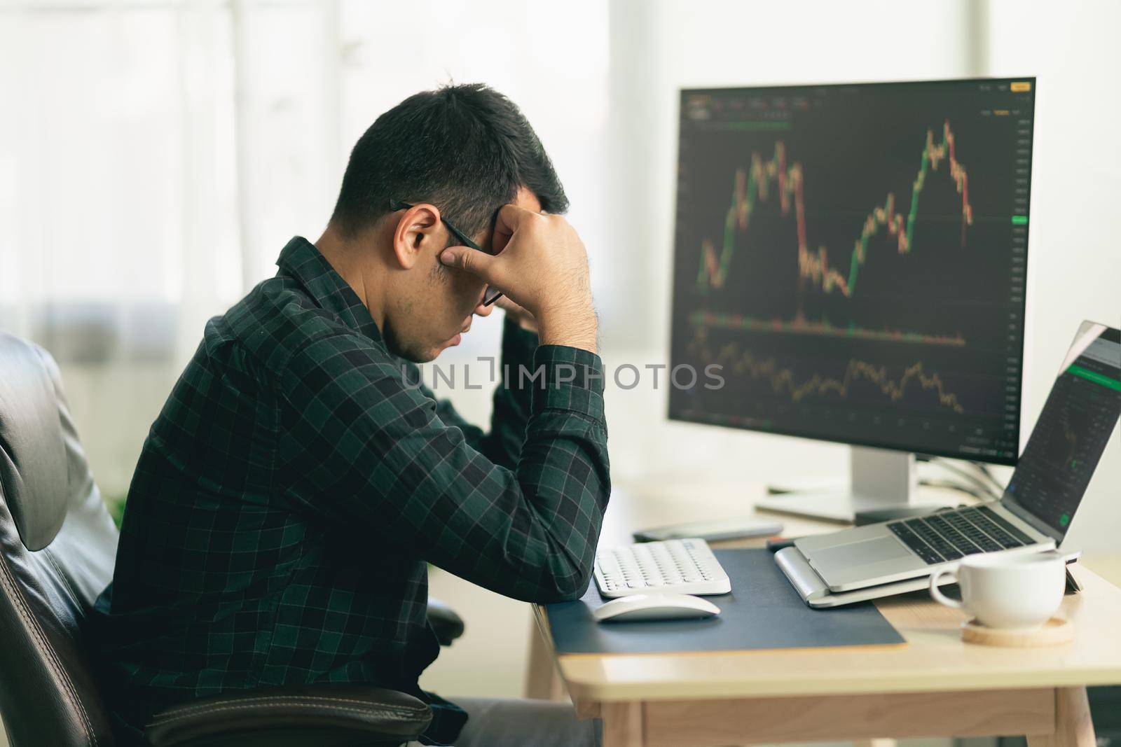 Asian man trader unhappy and serious sitting at home office in front of monitors with cryptocurrency graph holding smartphone monitoring cryptocurrency price by Wmpix