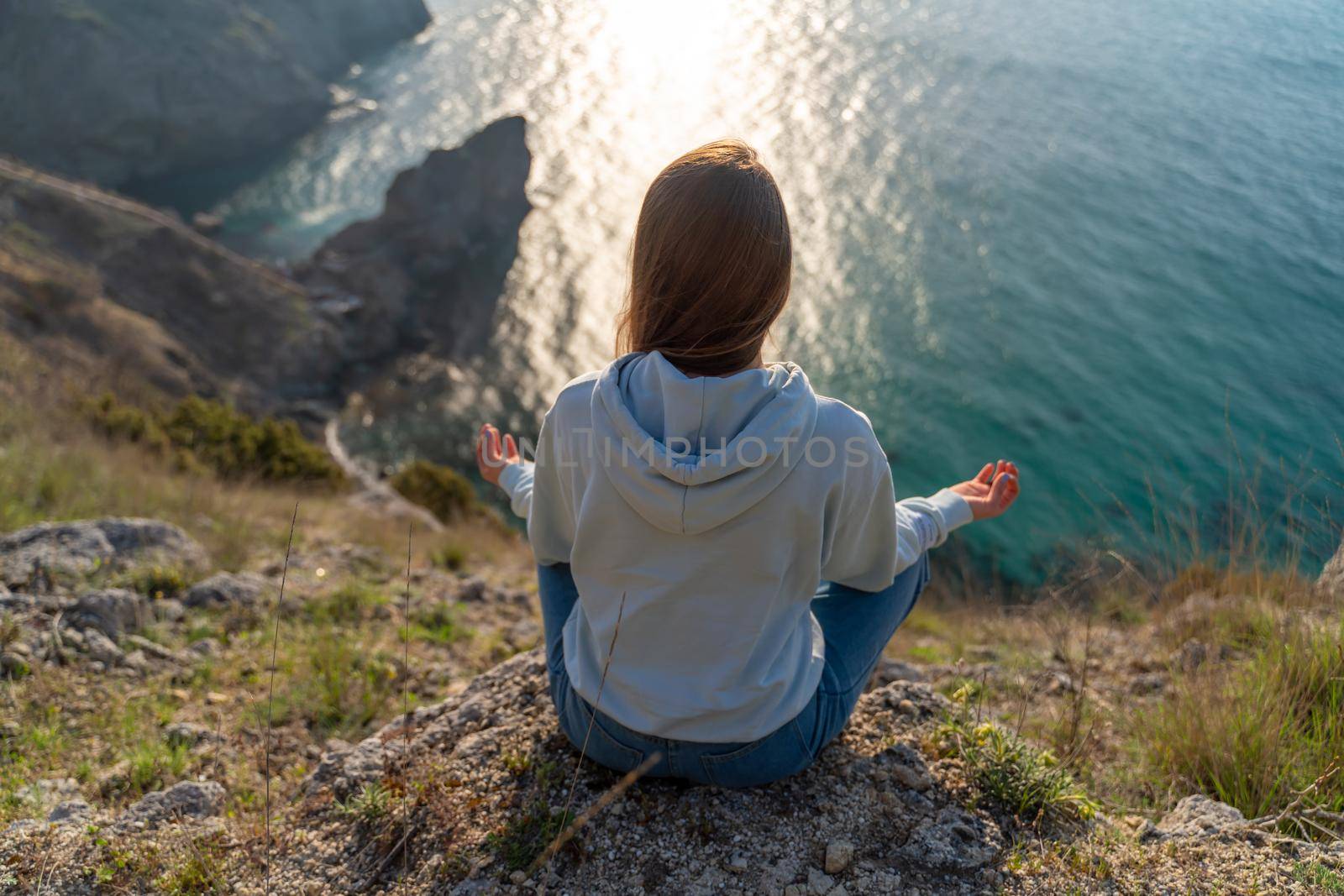 Woman tourist enjoying the sunset over the sea mountain landscape. Sits outdoors on a rock above the sea. She is wearing jeans and a blue hoodie. Healthy lifestyle, harmony and meditation by Matiunina