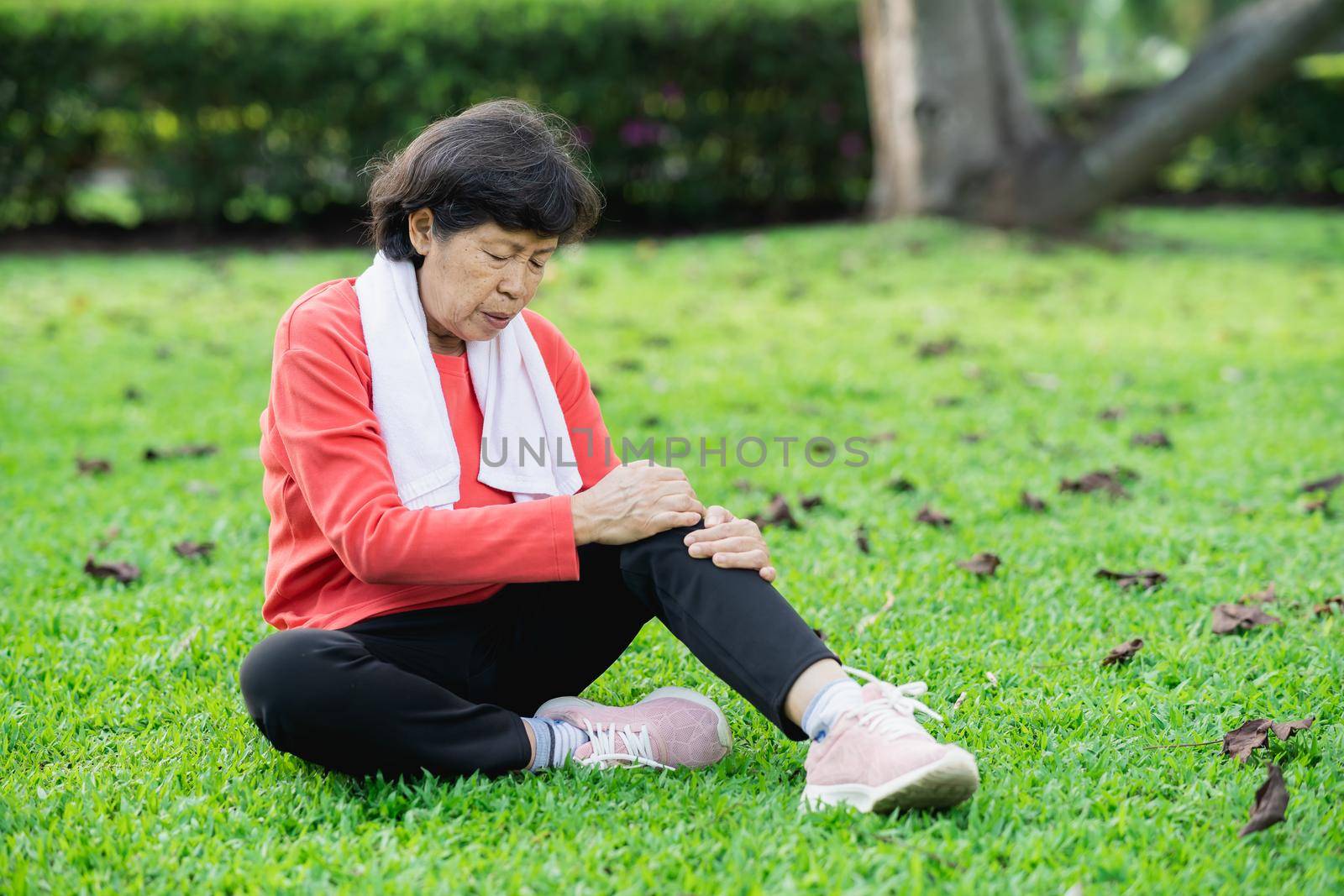 Senior asian woman with knee ankle pain while running in park. Senior asian woman sitting on the ground and holding painful knee. by Wmpix