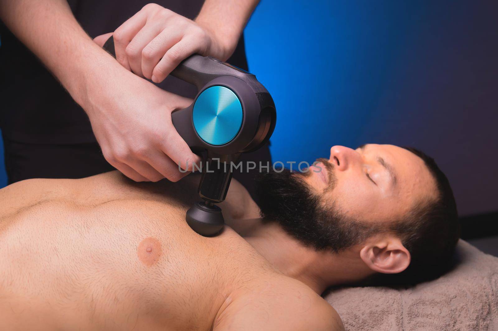 Medium shot of caucasian professional male massage therapist getting pectoral muscles with massage gun percussion tool of muscular athlete, in spa treatments, lying on back in massage table. by yanik88