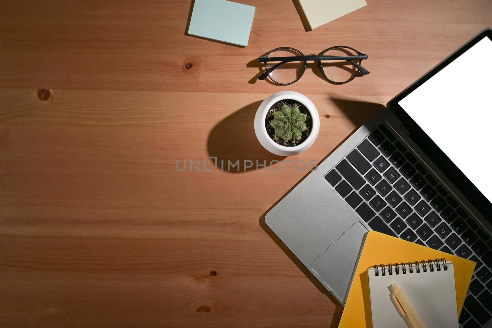 Flat lay of office desk with laptop, notepad, glasses, coffee cup and sticky notes on wooded table.