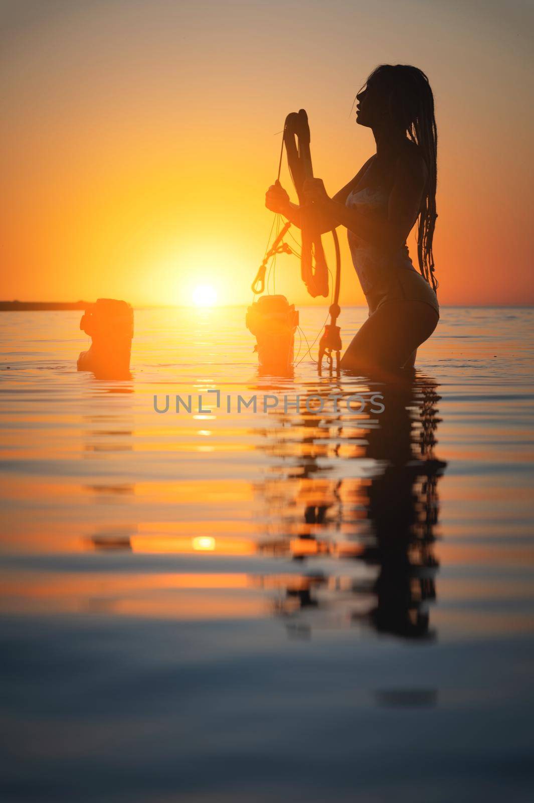 Silhouette frame. A young slim sexy female kitesurfer with a plank and a kiteboard stands in the water in the shallow water at sunset. Water sports. Stylized frame by yanik88