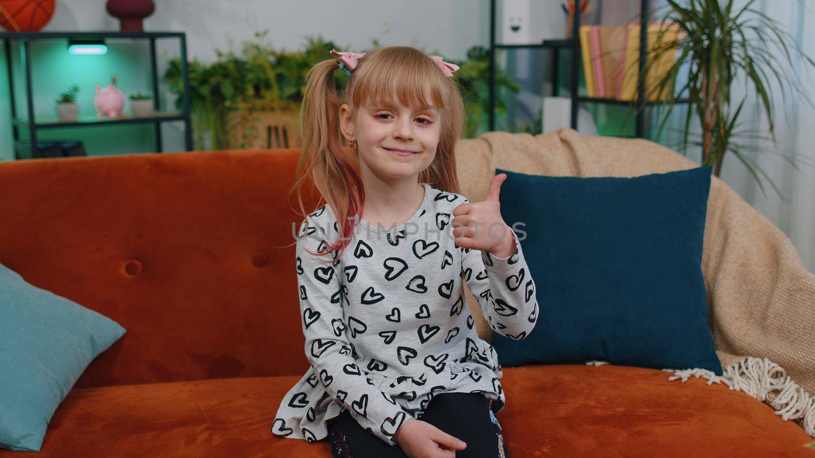 Happy young little children girl raises thumbs up agrees with something or gives positive reply recommends advertisement likes good feedback. Cheerful positive toddler child kid sits on sofa at home