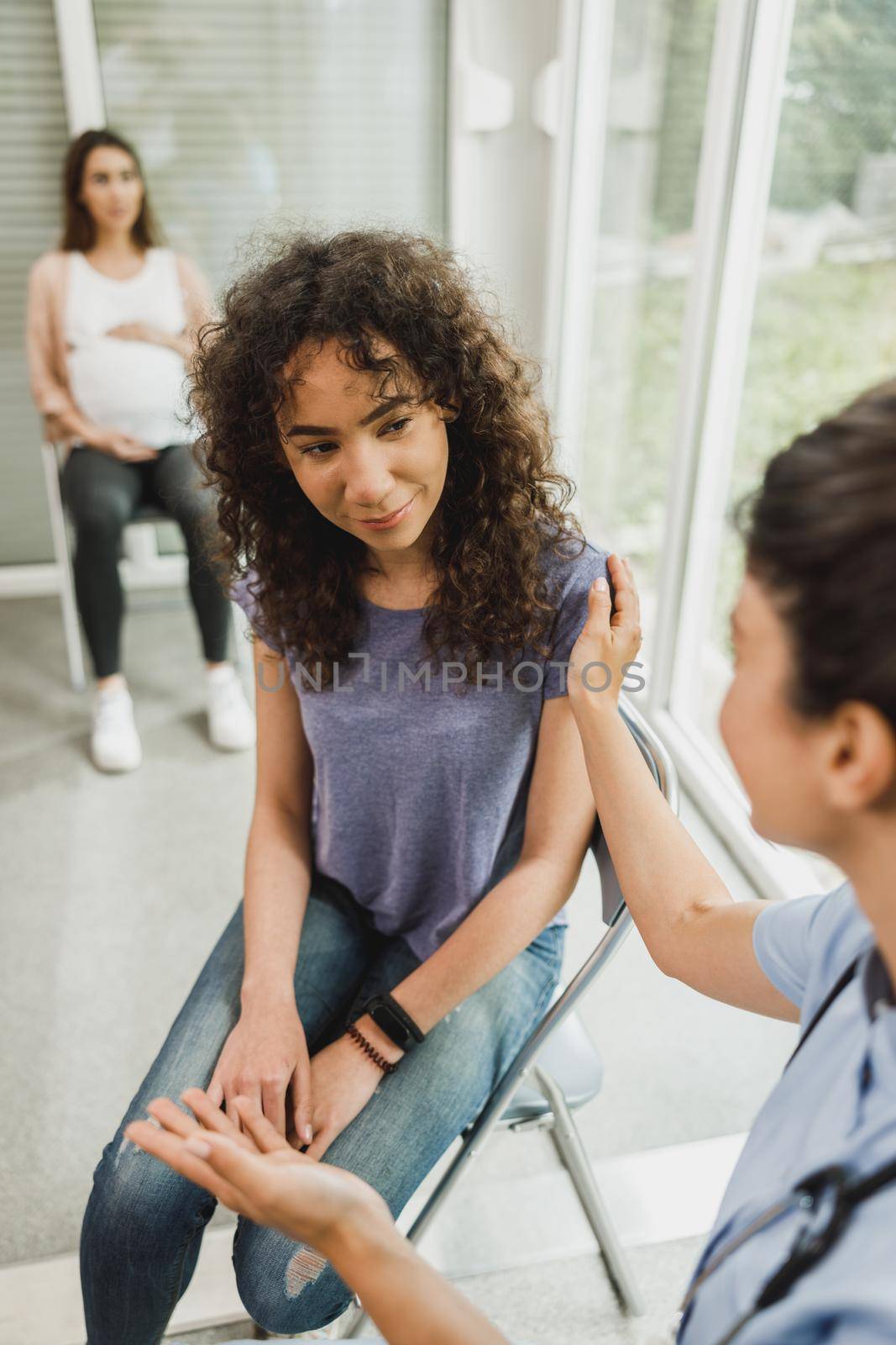 Upset African American teenage girl talks with nurse while waits for gynecologist check up.