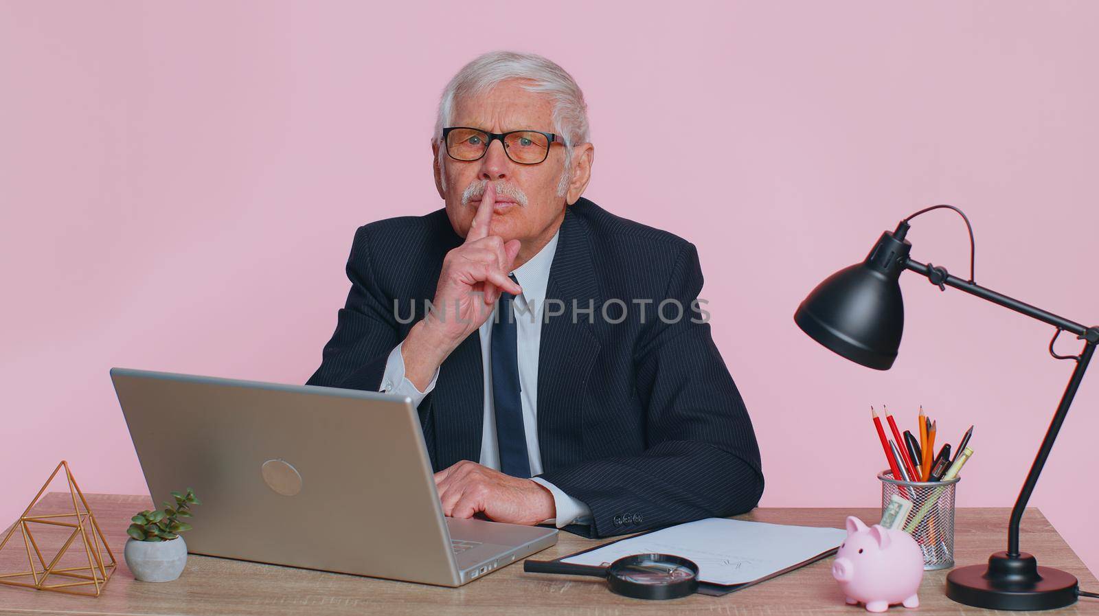 Shh be quiet please. Senior businessman in suit presses index finger to lips makes silence gesture sign do not tells secret sits at workplace desk at office. Elderly man on pink studio wall background