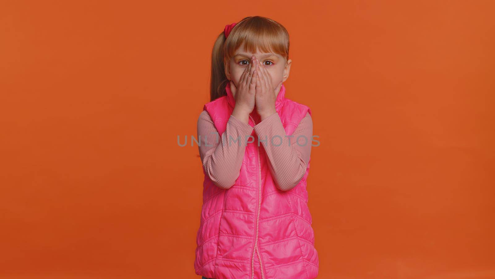 Oh my God, Wow. Excited amazed winner girl raising hands in surprise looking at camera with big eyes, shocked by sudden victory. Sincere young little child kid isolated on orange studio background