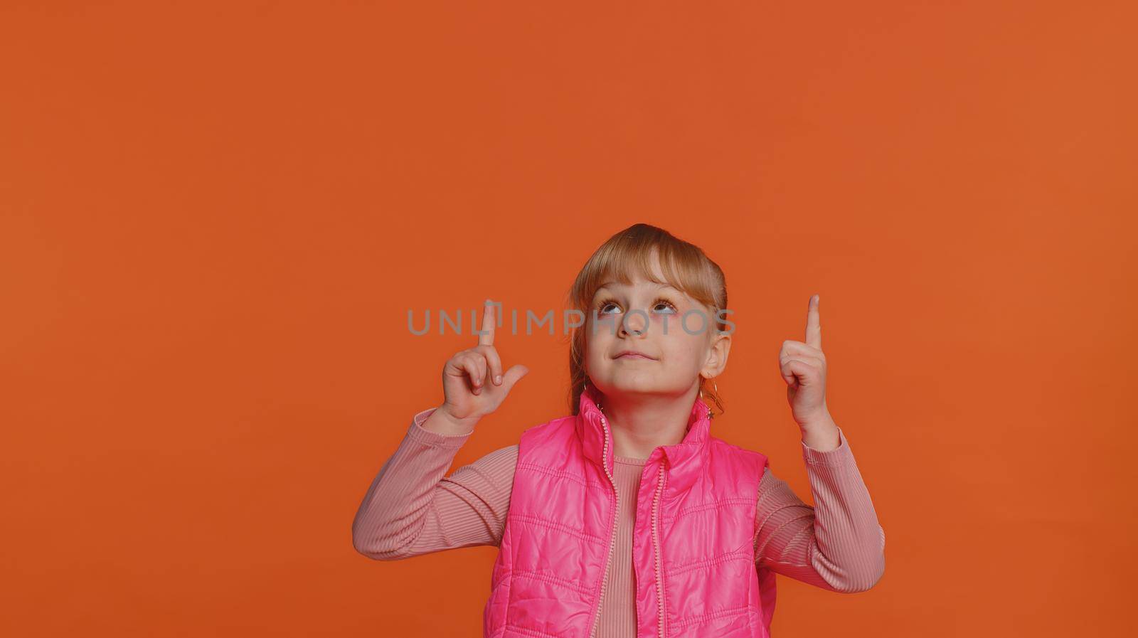 Toddler children girl showing thumbs up, pointing up empty place, advertising area for commercial text, copy space for goods promotion. Young one little child kid isolated on orange studio background